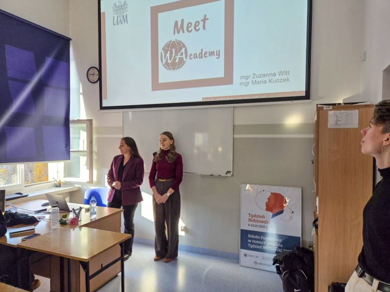 zdjęcie: dwie osoby stoją w sali lekcyjnej, trzecia stoi z boku i przygląda się im;p za nimi, na ekranie, slajd z prezentacji