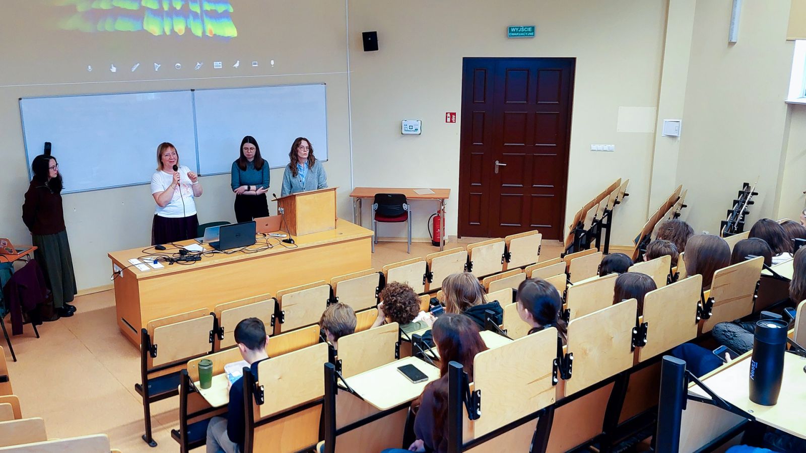 zdjęcie: trzy osoby stoją w sali wykładowej; na ekranie za nimi: obraz z rzutnika; przed nimi: zgromadzona w sali publiczność