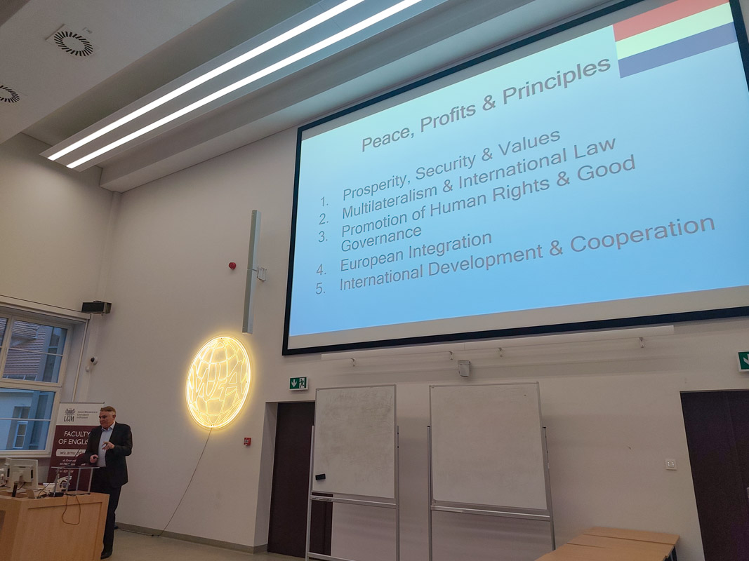 zdjęcie: osoba stoi za mównicą w sali wykładowej; na ścianie za nią: ekran ze slajdem z prezentacji mutlimedialnej