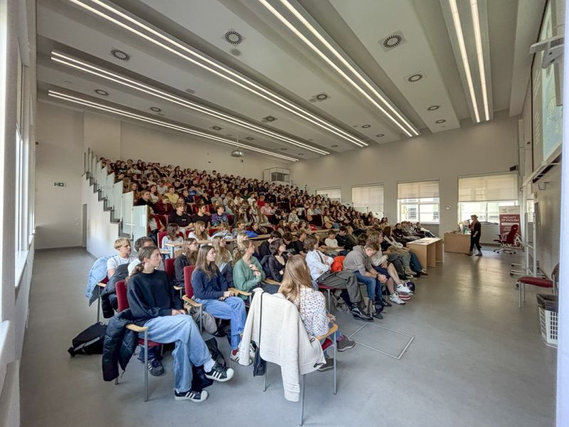 Duża grupa uczniów i uczennic uczestnicząca w wykładzie w przestronnej auli, słuchająca prezentacji prowadzącej przy ekranie.