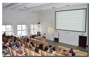 photograph: two people stand behind a table in the lecture hall; behind them on the screen is a slide from a multimedia presentation; in front of them is the audience gathered in the hall