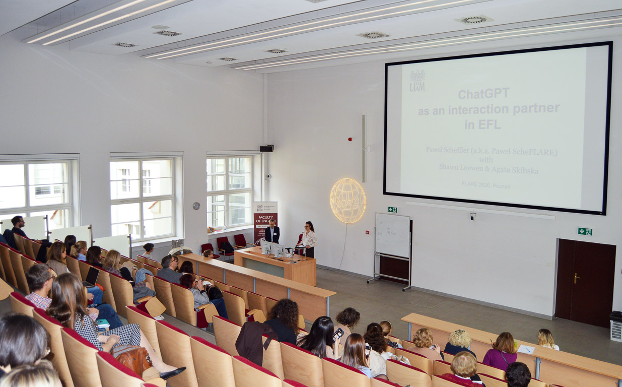 photograph: two people stand behind a table in the lecture hall; behind them on the screen is a slide from a multimedia presentation; in front of them is the audience gathered in the hall