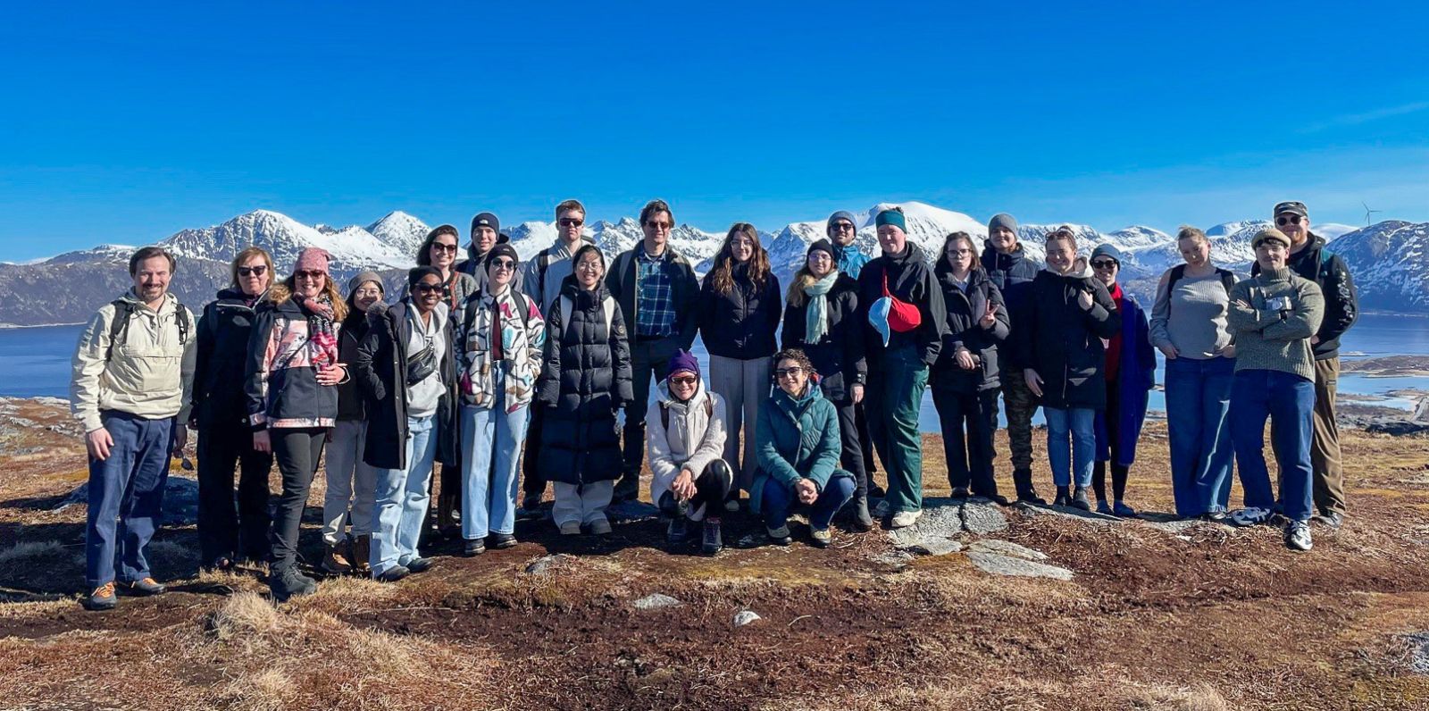photograph: twenty-four people by a rock; backround: a large pool of water, rocks, a blue sky