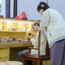 photograph: one person sits at, another stands close to a desk with pottery materials