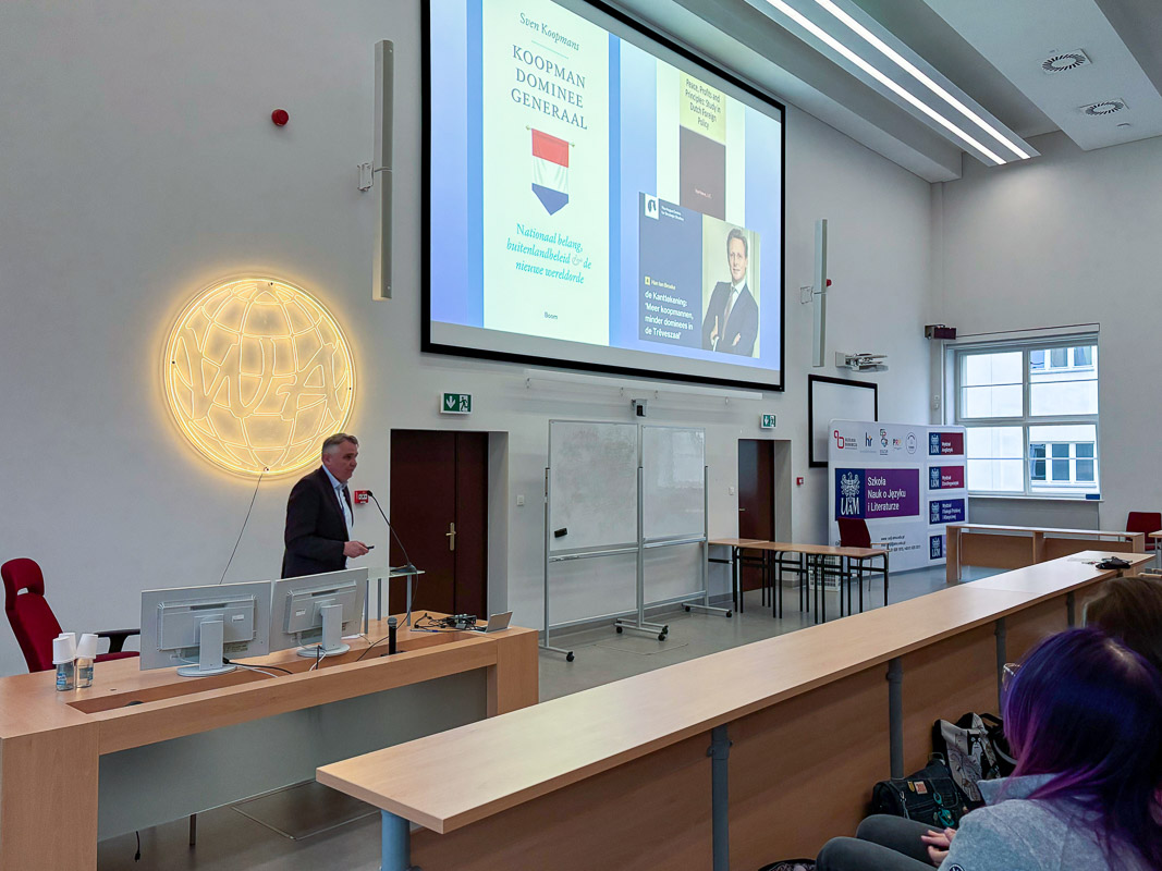 zdjęcie: osoba stoi za mównicą w sali wykładowej; na ścianie za nią: ekran ze slajdem z prezentacji mutlimedialnej; przed nią: publiczność