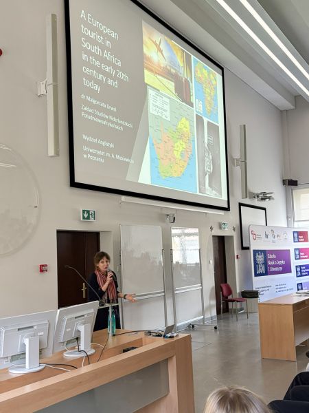 Osoba prowadząca prezentację w auli, stojąca przy mównicy i omawiająca slajd dotyczący tematyki podróży europejskiego turysty w RPA.