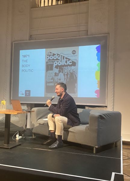 Marcin Markowicz siedzi na kanapie po prawej strony sceny i mówi do mikrofonu. Uśmiecha się. Za nim prezentacja a na niej okładka kanadyjskiego czasopisma i napis 1971 The Body Politic.