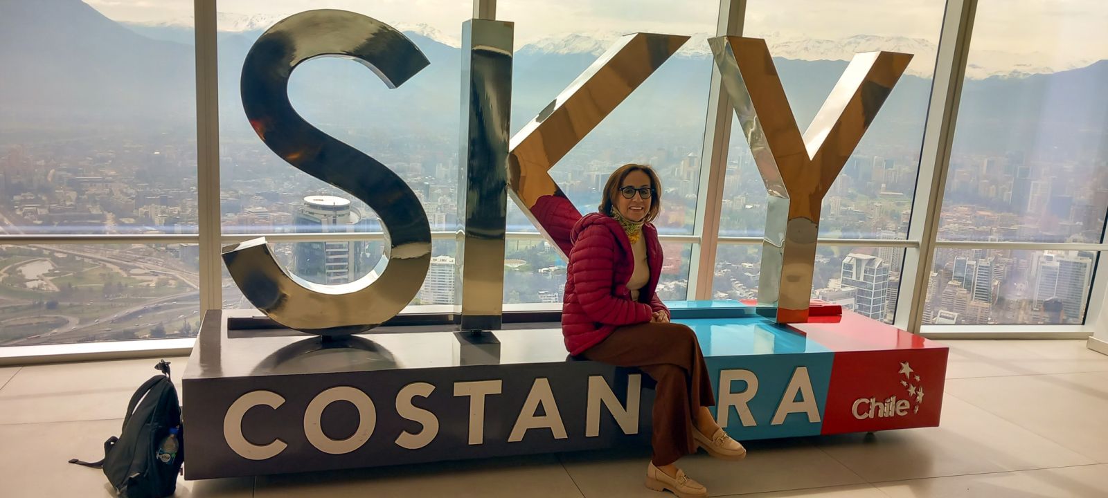 photograph: a person poses for a photograph at a bench next to a large window at a high landing of a tall bulding; behind them: a cityscape