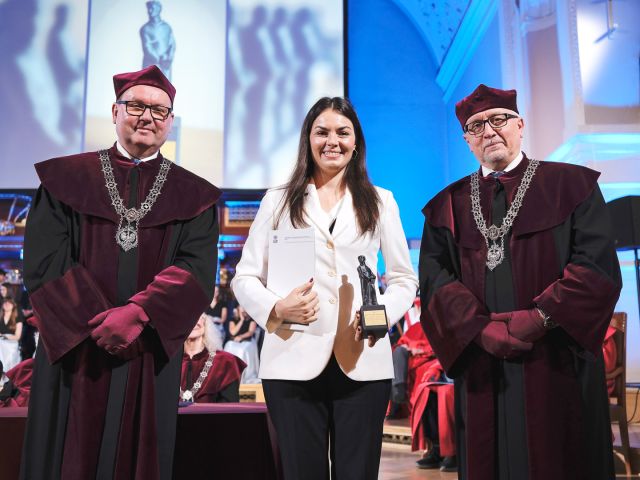 photograph: three persons pose for a photograph; two of them are wearing ceremonial robes; the third is holding a diploma and a statuette