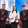 photograph: three persons pose for a photograph; two of them are wearing ceremonial robes; the third is holding a diploma and a statuette