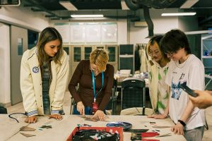 photograph: four people stand by a desk with materials for making glass jewelry