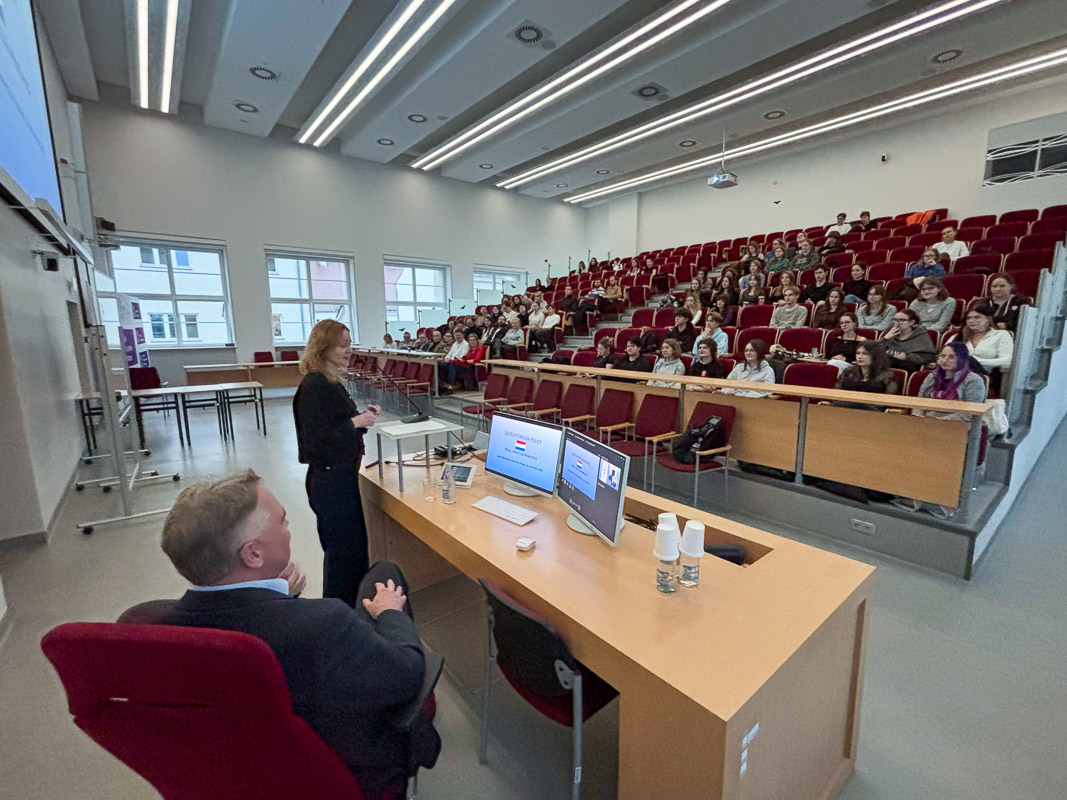 zdjęcie: jedna osoba stoi, druga siedzi za mównicą w sali wykładowej; przed nimi: publiczność siedząca w kilku rzędach foteli