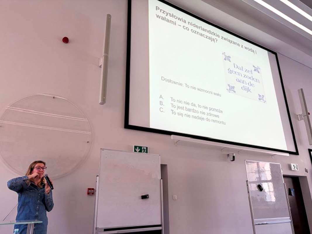 Osoba prowadząca zajęcia stoi przy mównicy i omawia slajd o niderlandzkich przysłowiach związanych z wodą, wyświetlony na dużym ekranie w auli.