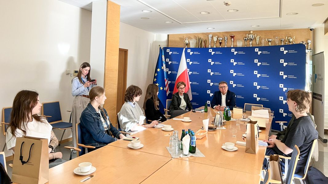 zdjęcie: siedem osób siedzi wokół stołu w sali konferencyjnej, jedna osoba stoi dalej; osoby przy stole rozmawiają