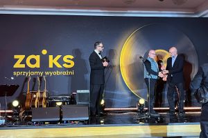 zdjęcie: scena, na niej trzy osoby, pierwsza z prawej wręcza statuetkę osobie w środku kadru