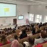 zdjęcie: osoba stojąca w sali wykładowej; na ekranie za nią: obraz z rzutnika; przed nią: zgromadzona w sali publiczność