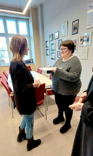 dwie osoby stojące przy stole konferencyjnym, jedna wręczająca drugiej dyplom gratulacyjny