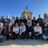 photograph: a group of nineteen people posing for a photograph, arranged into two rows: those behind standing, those in front squatting; in the background an element of architecture suggests an exterior location