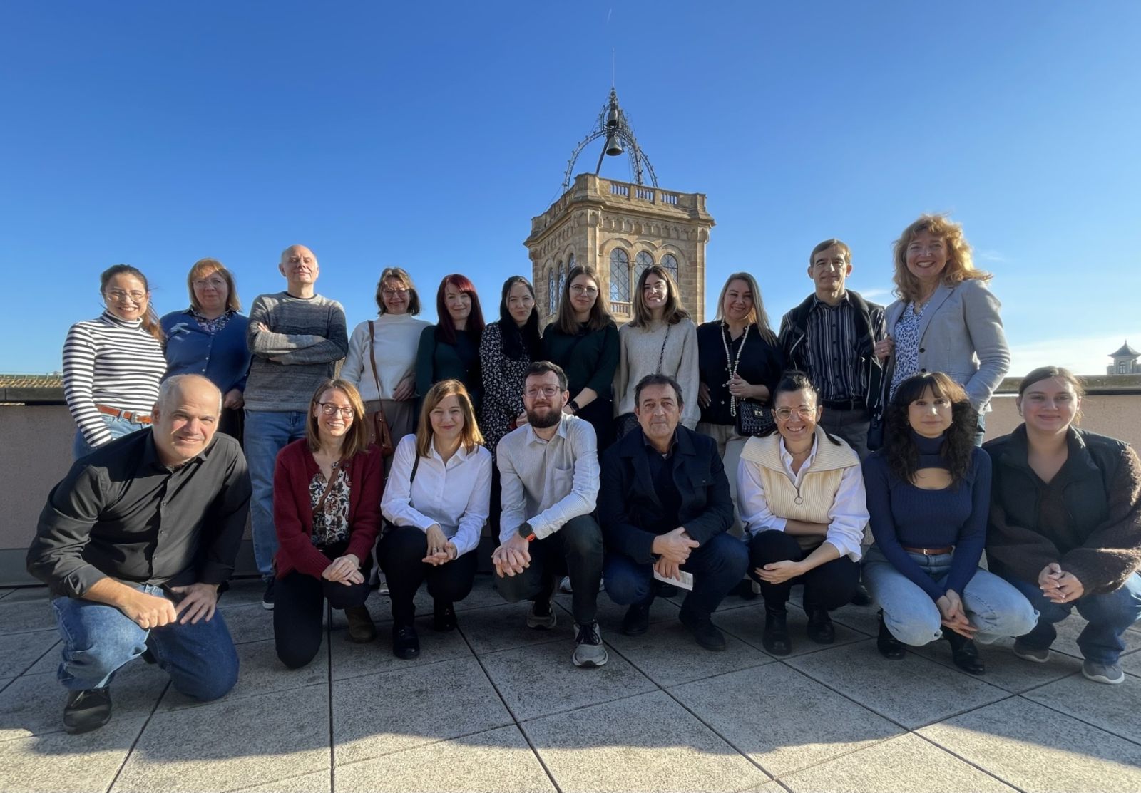 photograph: a group of nineteen people posing for a photograph, arranged into two rows: those behind standing, those in front squatting; in the background an element of architecture suggests an exterior location