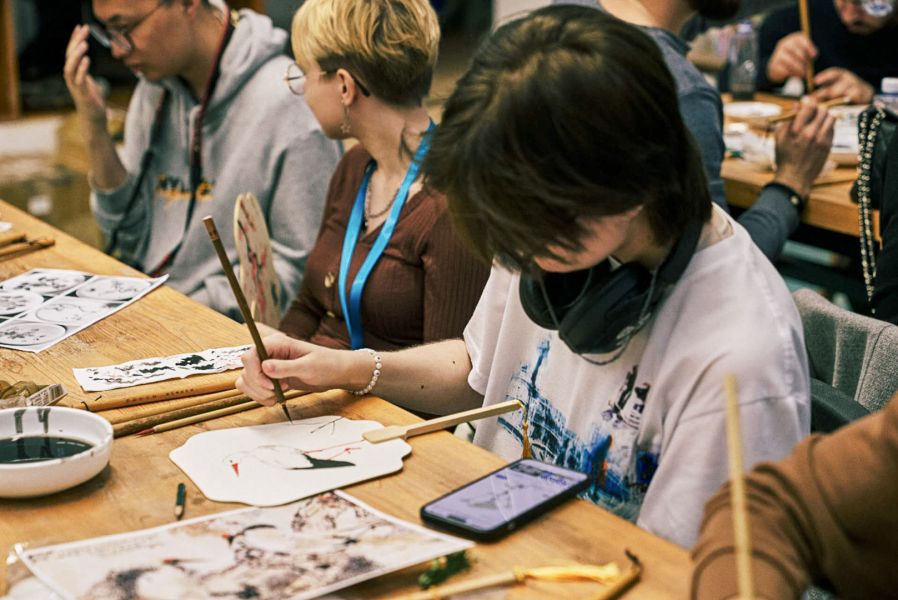 photograph: a person sits at a desk and decorates a paper-and-wood fan; two more persons sitting by the same desk are also in the frame