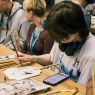 photograph: a person sits at a desk and decorates a paper-and-wood fan; two more persons sitting by the same desk are also in the frame