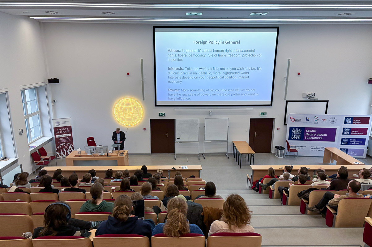 zdjęcie: osoba stoi za mównicą w sali wykładowej; na ścianie za nią: ekran ze slajdem z prezentacji mutlimedialnej; przed nią: publiczność siedząca w kilku rzędach foteli