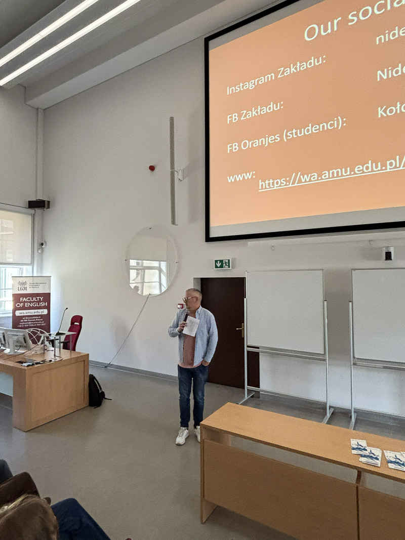 Osoba prowadząca stoi w auli z mikrofonem, omawiając slajd z informacjami o kanałach społecznościowych Zakładu, wyświetlony na dużym ekranie.