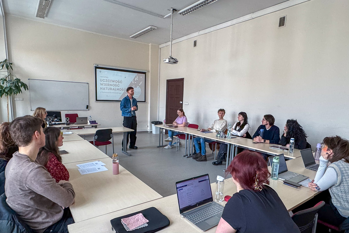 zdjęcie: jedna osoba przemawia, stojąc przed ekranem z wyświetlonym slajdem z prezentacji; kilkanaście osób siedzi za stołami ustawionymi dookoła osoby przemiawiającej