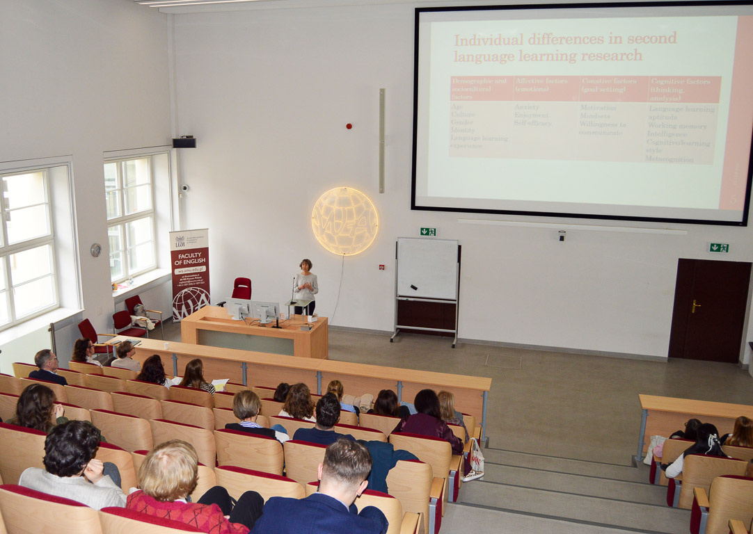 photograph: a person stands behind a table in a lecture hall; a slide from a multimedia presentation is displayed on a screen behind them