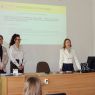 zdjęcie: trzy osoby stoją za stołem w sali seminaryjnej; za nimi na ekranie slajd z prezentacji multimedialnej