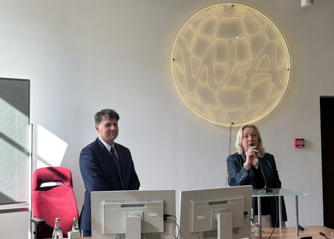 zdjęcie: sala wykładowa: za mównicą stoją dwie osoby: jedna przemawia do mikrofonu, druga patrzy w kadr, uśmiechając się