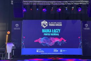 photograph: scene flooded with saturated blueish light; on the screen behind the scene: captions in Polish