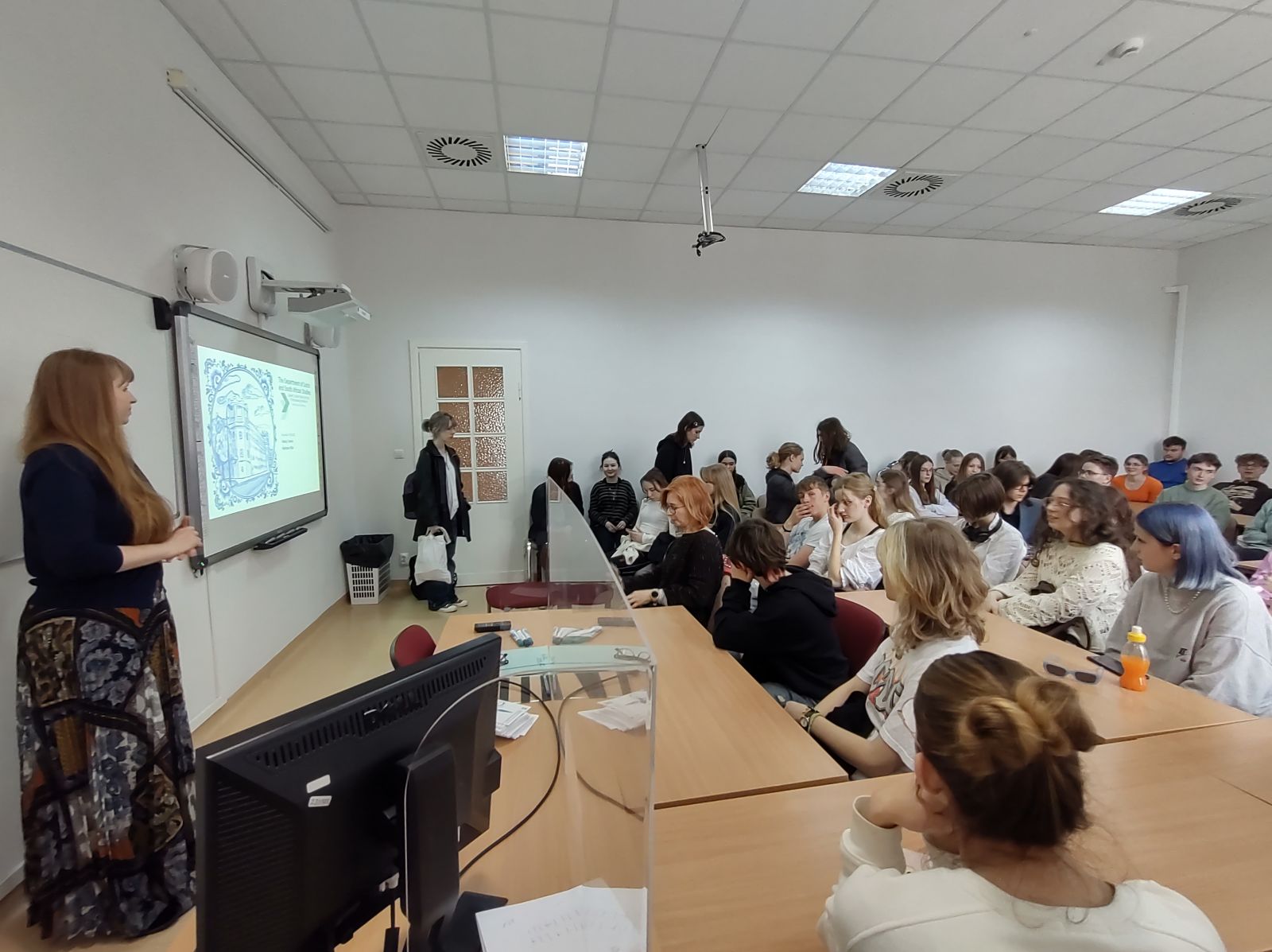 zdjęcie: osoba stojąca w sali wykładowej; na ekranie za nią: obraz z rzutnika; przed nią: zgromadzona w sali publiczność