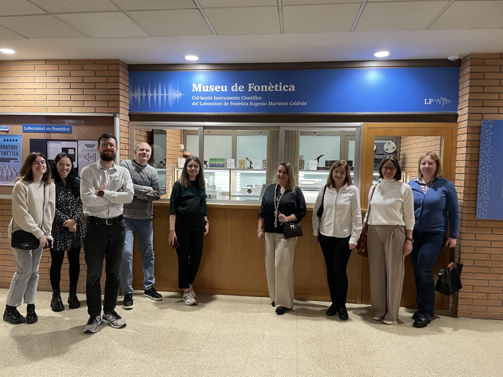 photograph: a group of nine people posing for a photograph in a building hall, behind them is a display with the caption in Catalan {Museu de Fonètica}