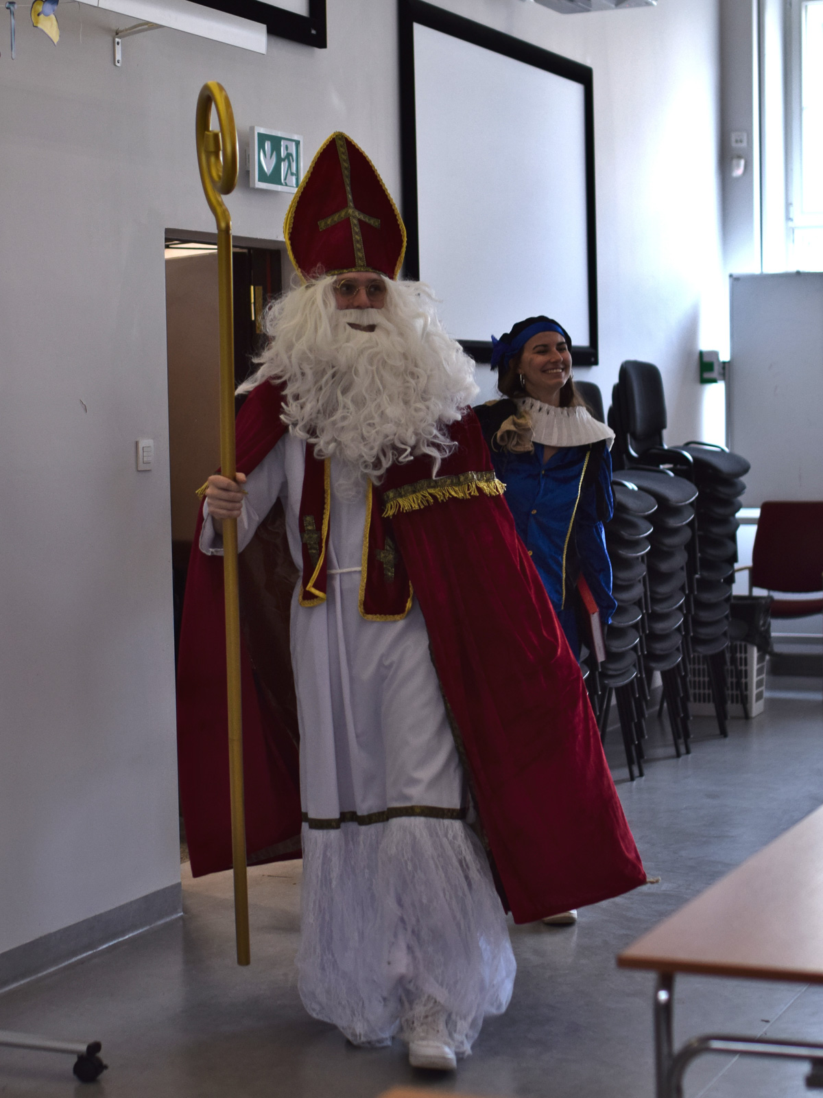 zdjęcie: osoba w przebraniu Sinterklaasa wchodzi do sali, za nią widoczna osobu przebrana za postać Zwarte Piet