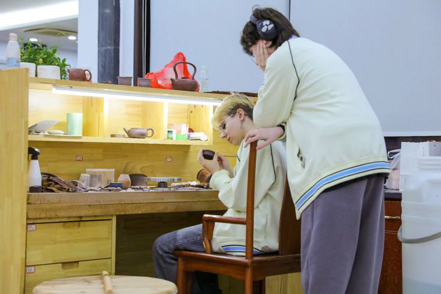 zdjęcie: jedna osoba siedzi, druga osoba stoi przy stole z materiałami garncarskimi