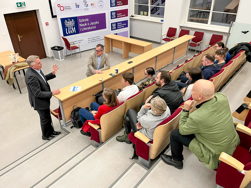 zdjęcie: grupa osób dyskutuje w sali wykładowej: dwie osoby stoją, pozostałych dziesięcioro siedzi w rzędach