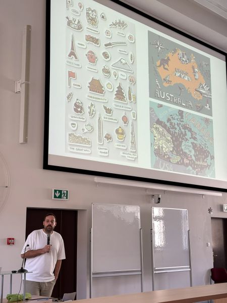 Osoba prowadząca stoi w auli z mikrofonem, omawiając slajd przedstawiający ilustracje i mapy różnych regionów świata.