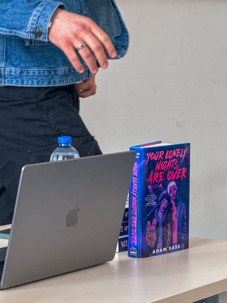 zdjęcie: osoba stoi za stołem, na którym znajdują się: laptop i książka w twardej oprawie: Adam Sass „Your Lonely Nights Are Over”