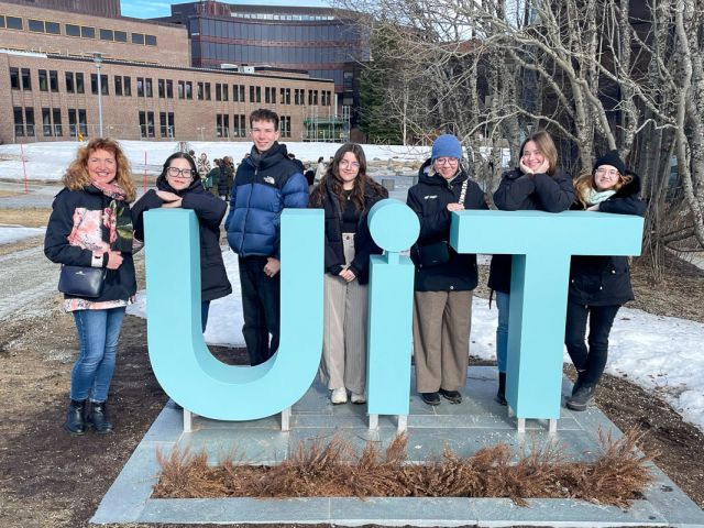 photograph: seven people pose by a monument made of the letters {U}, {i}, and {T}; a buliding and a clear sky in the background