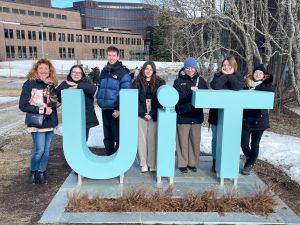 photograph: seven people pose by a monument made of the letters {U}, {i}, and {T}; a buliding and a clear sky in the background