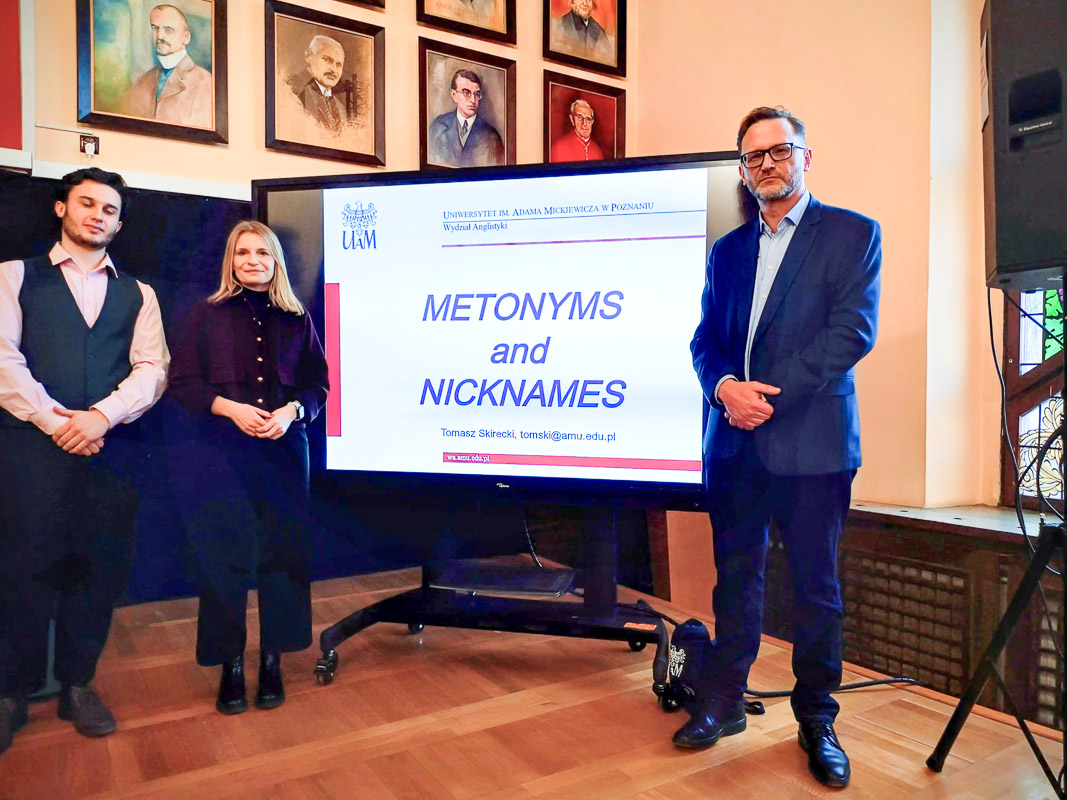 zdjęcie: trzy osoby stoją na podium przy ekranie, na którym wyświetlany jest slajd z tytułem przentacja „Metonyms and Nicknames”