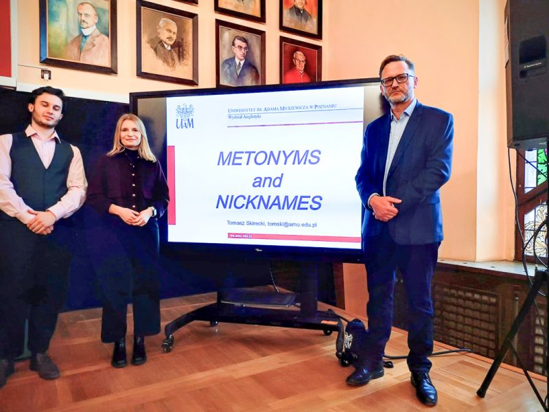 zdjęcie: trzy osoby stoją na podium przy ekranie, na którym wyświetlany jest slajd z tytułem przentacja „Metonyms and Nicknames”