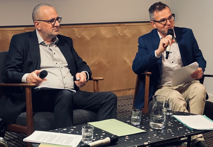 zdjęcie: dwie osoby siedzą z mikrofonami podczas rozmowy na scenie, przy stole z notatkami i szklankami