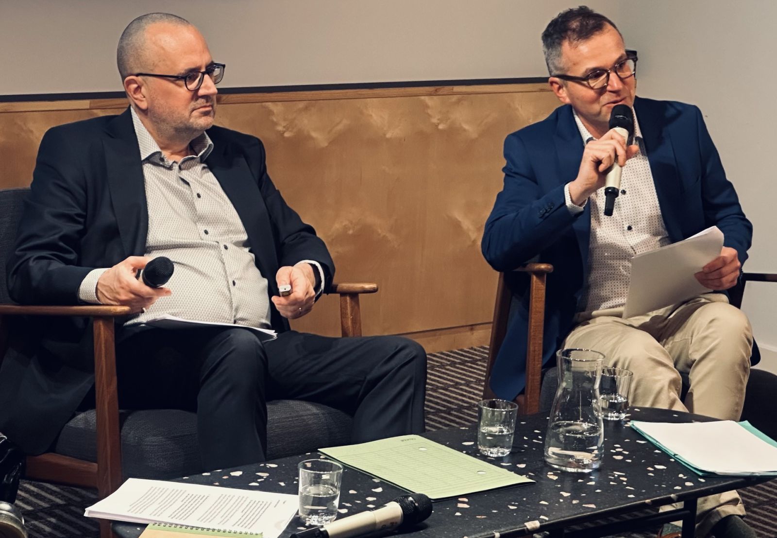 zdjęcie: dwie osoby siedzą z mikrofonami podczas rozmowy na scenie, przy stole z notatkami i szklankami