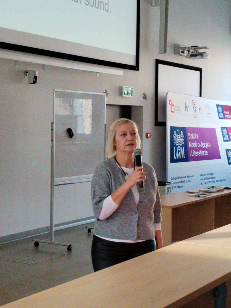 Osoba prowadząca stoi w auli z mikrofonem, mówiąc do publiczności; obok widoczny jest ekran z informacjami o Szkole Nauk o Języku i Literaturze UAM.