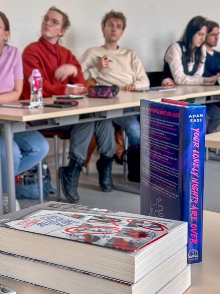 zdjęcie: stół, na nim: książka w twardej oprawie: Adam Sass „Your Lonely Nights Are Over” oraz dwie inne, niezidentyfikowane książki; w tle, w pewnej odległości: kilka osób siedzących za innym stołem