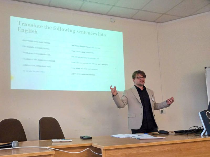 zdjęcie: osoba stoi za stołem w sali seminaryjnej; za nią na ekranie slajd z prezentacji multimedialnej