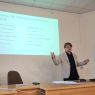 zdjęcie: osoba stoi za stołem w sali seminaryjnej; za nią na ekranie slajd z prezentacji multimedialnej