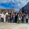 photograph: twenty-six people pose in an open space; some bulidings and a clear sky in the background